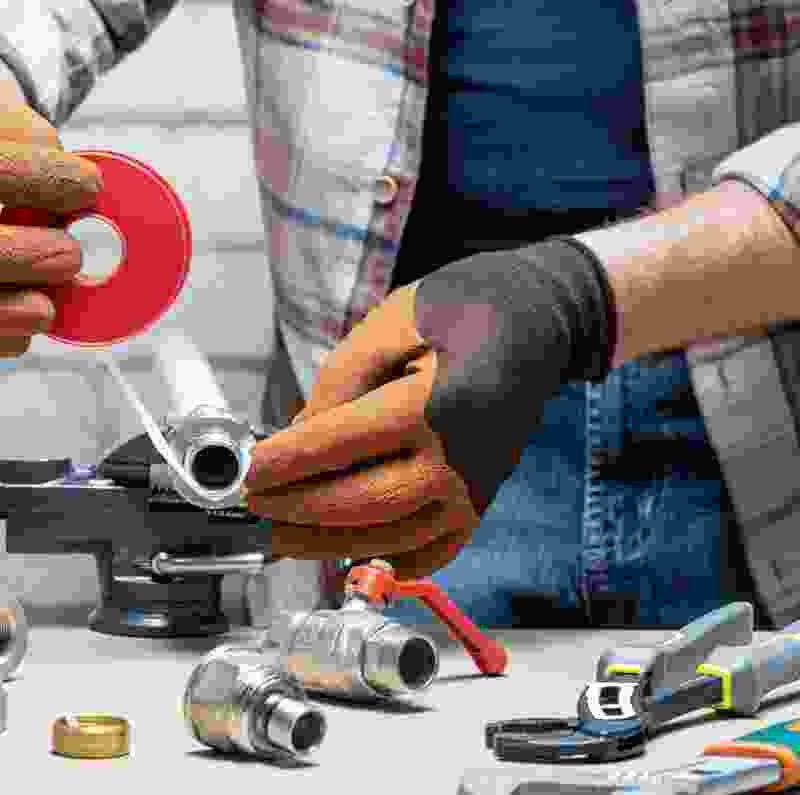 Person applying plumbing tape to a metal pipe fitting, surrounded by various plumbing tools and connectors, illustrating handyman services offered by Extra Electrical.