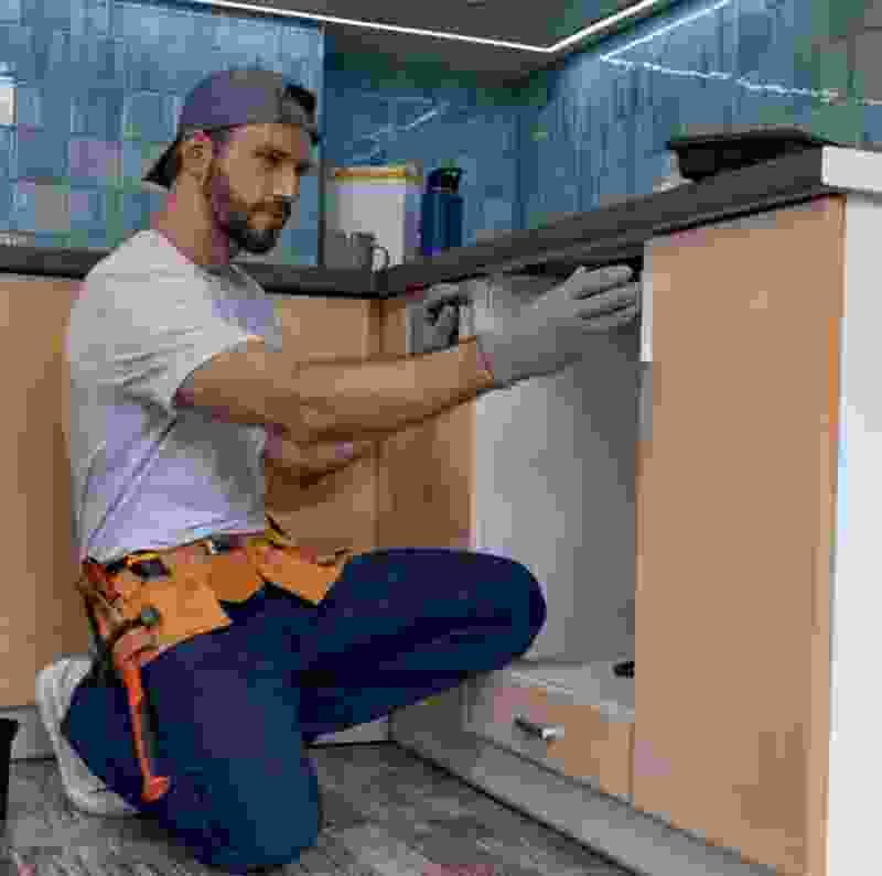 Man in work attire kneeling and inspecting kitchen cabinetry, showcasing handyman services for commercial eco-friendly pest control solutions.